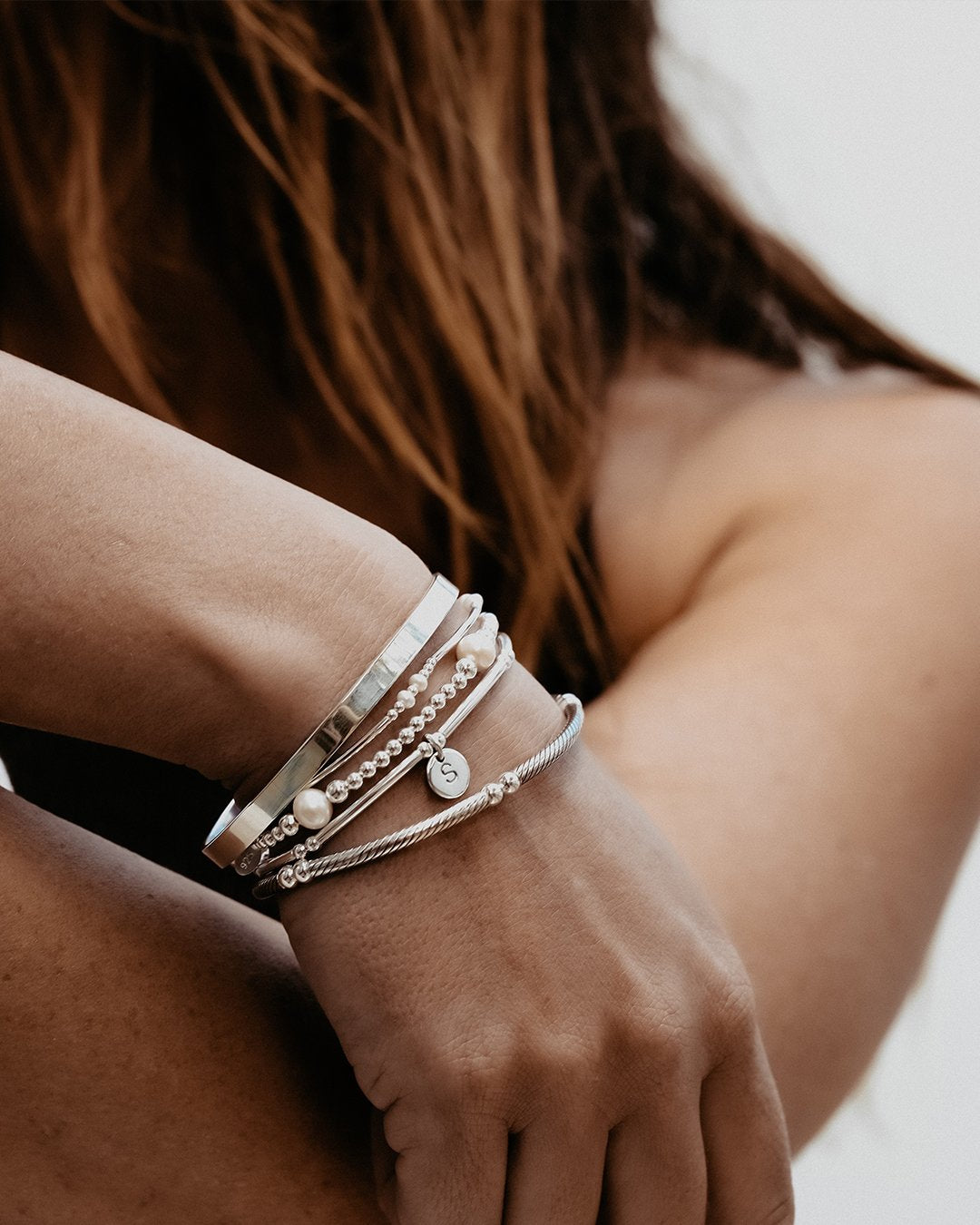 A person's forearm wearing a stack of sterling silver bracelets featuring different designs, including a plain cuff, twisted bar, large pearl, two pearl, and a love letter bracelet. Jewellery by Petals Australia